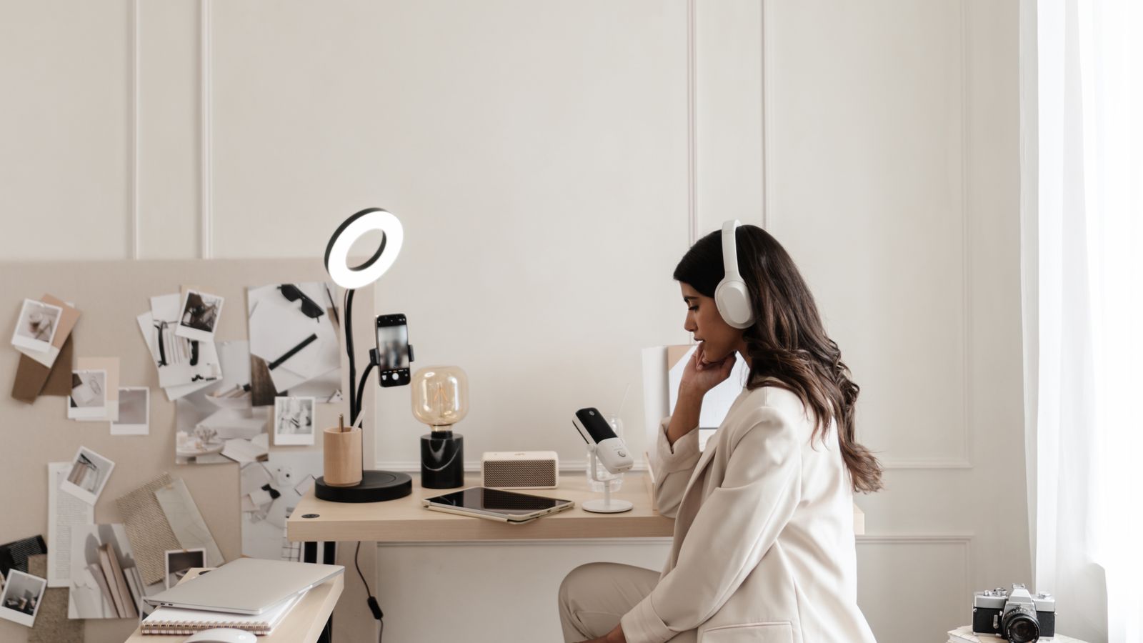 A woman in headphones at a desk with a phone on a ring light stand and a mood board of photos behind her