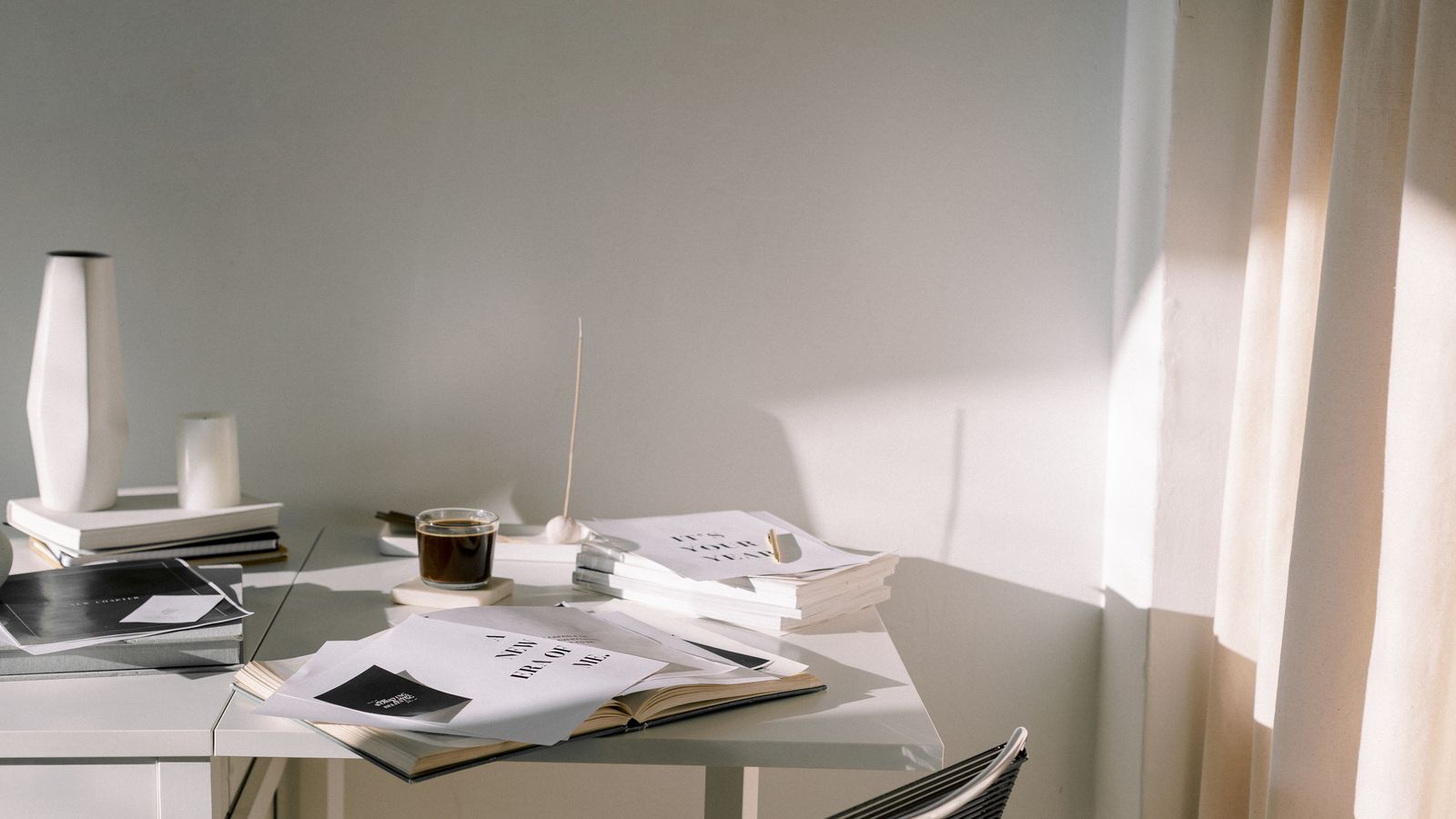 Loose papers and an open book on a white desk in soft morning light, beside a sheer curtain