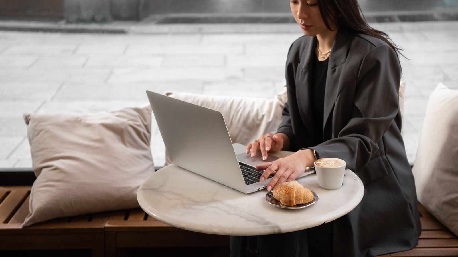 A woman in a dark blazer typing on a laptop at a small marble café table, with a cappuccino and a croissant