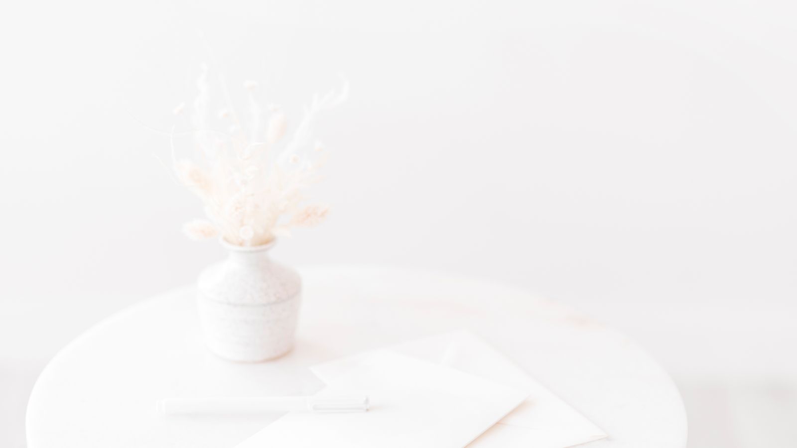 A white envelope and a pen on a round marble table beside a vase of dried flowers